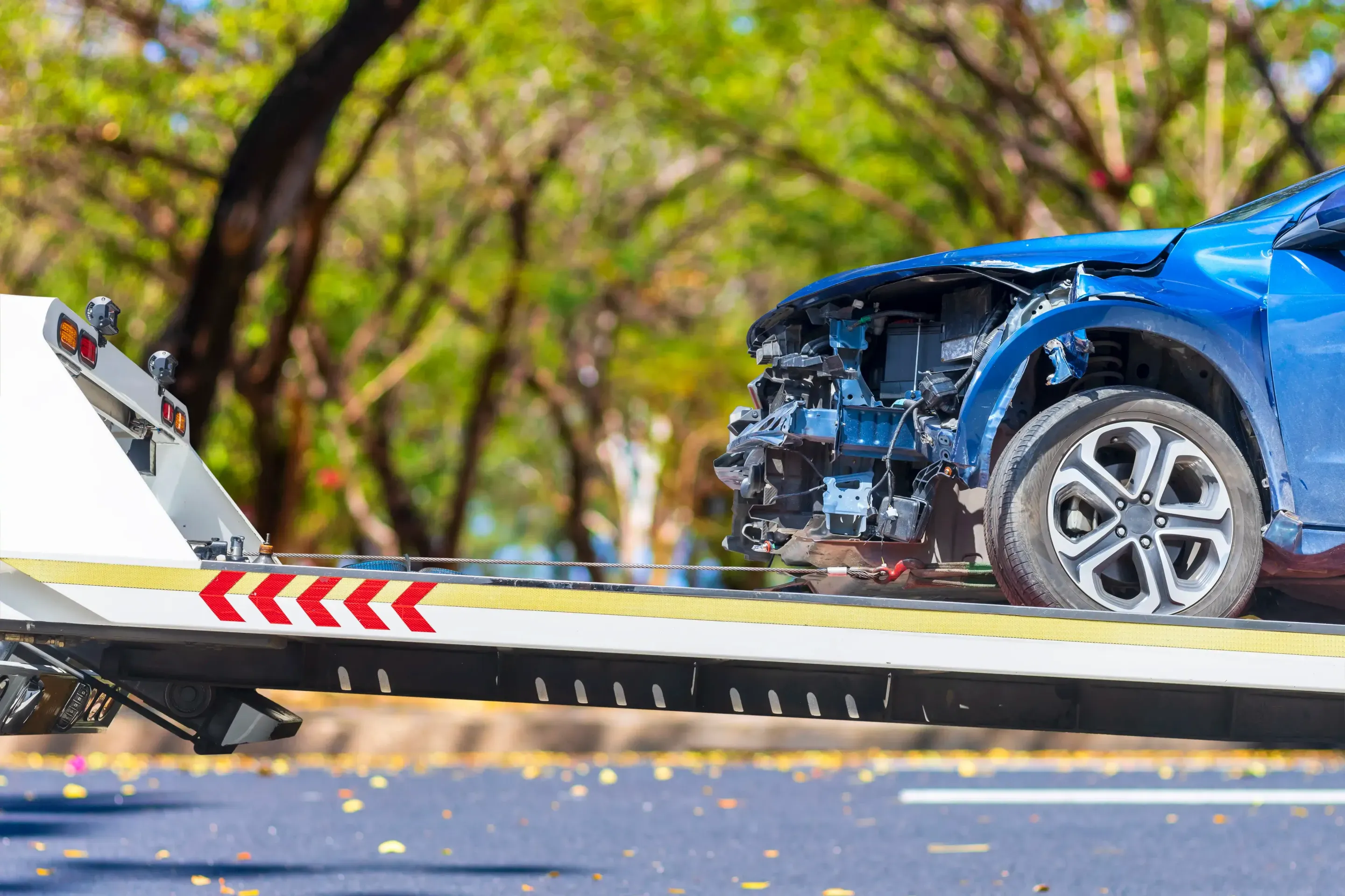 Damaged blue car with a crushed front end strapped onto a white flatbed on a tree-lined road